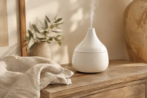 Diffuseur électrique blanc en céramique posé sur du bois patiné, avec un textile en lin et une touche végétale.